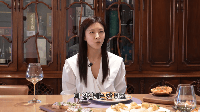 Ha Ji-won  in a white blouse speaks while seated at a table with various food items and a glass of wine, surrounded by wooden cabinetry.
