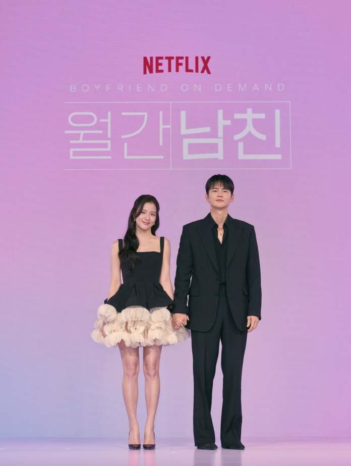 Jisoo and Seo In-guk holding hands at a promotional event for 'Boyfriend on Demand' by Netflix. Jisoo  is wearing a short black dress with a ruffled beige hem, while Seo In-guk is dressed in a black pinstripe suit.
