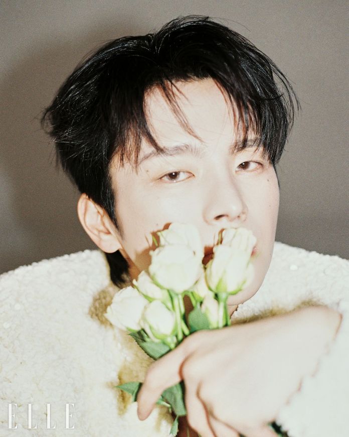 A close-up portrait of Seo In-guk with dark hair, holding a bouquet of white roses near their face, dressed in a textured white jacket, with a neutral background.