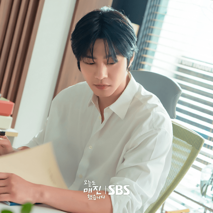Ahn hyo seop with dark, slightly wavy hair sits at a desk, looking thoughtfully at a document in his hands. He is wearing a white button-up shirt and is surrounded by a modern, well-lit office space with wooden accents and a chair in the background.