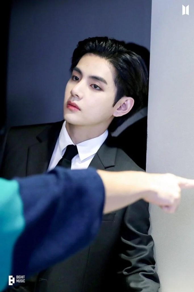 V in a black suit and tie leans against a wall, looking thoughtfully to the side, while a hand points towards him in the background.