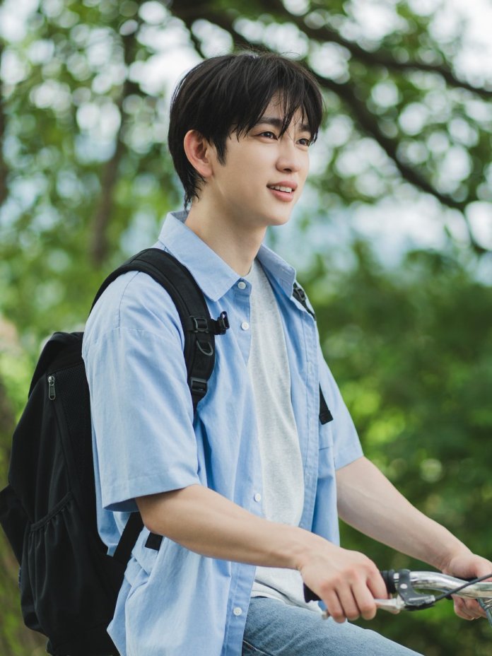 Park Jinyoung with dark hair smiles while sitting on a bicycle, wearing a light blue shirt and a gray t-shirt underneath. He has a black backpack on and is surrounded by greenery.