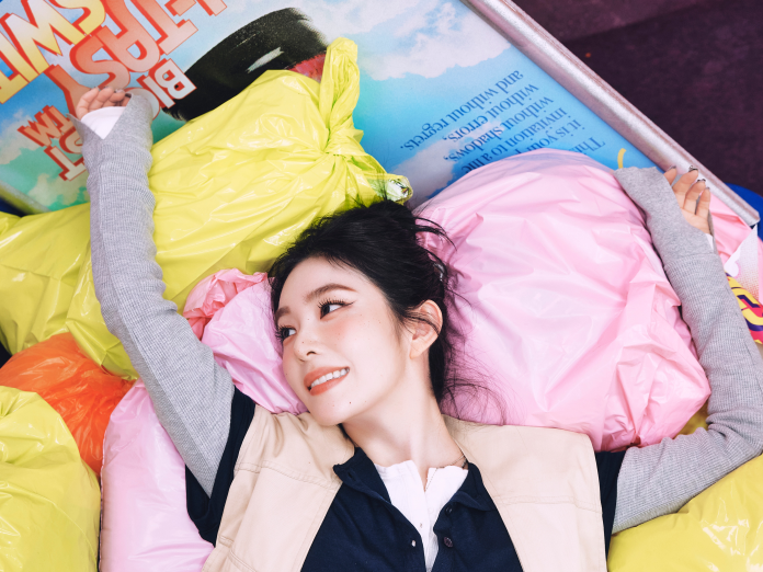 A smiling Irene lies among colorful plastic bags and a magazine, showcasing a playful and relaxed atmosphere.