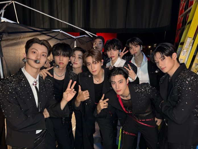 The boyz posing together backstage, wearing stylish black outfits adorned with sequins, some with microphones. They are smiling and making peace signs.