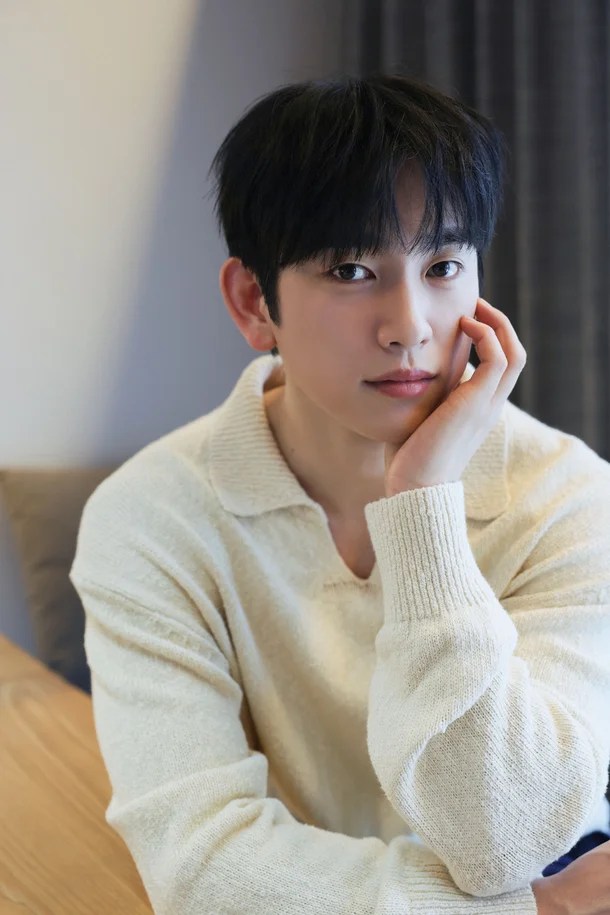 Park Jinyoung with short black hair wearing a light-colored sweater, resting their chin on their hand while sitting at a wooden table, with soft lighting and a neutral background.
