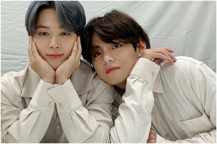V and Jimin posing together, one resting his chin on his hands and smiling, while the other leans in closely with a gentle expression. They are wearing matching light-colored shirts, with a soft backdrop.