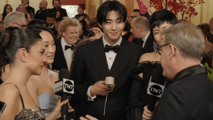 A stylish group of individuals, including Ahn Hyo-seop in a tuxedo and several women in elegant gowns, engage in conversation during a red carpet event, with cameras and onlookers in the background.