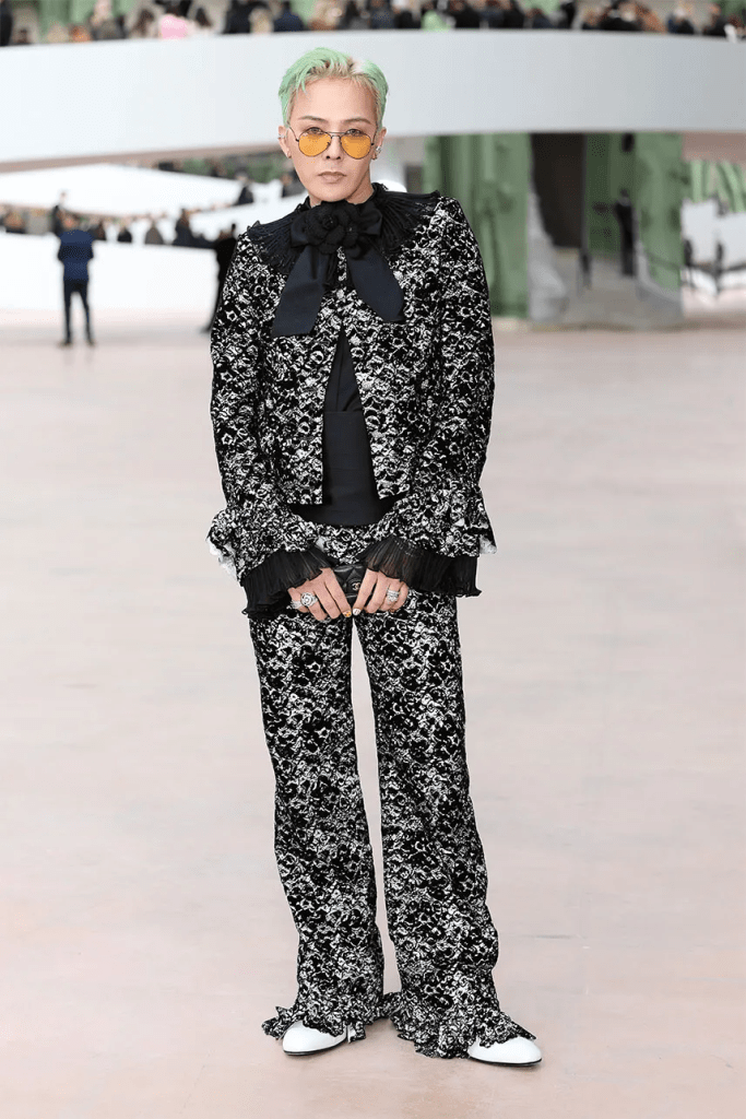 G-Dragon with short green hair wearing a black and white patterned suit with ruffled details and a large bow tie, accessorized with yellow sunglasses and bold rings, posing confidently in a modern outdoor setting.