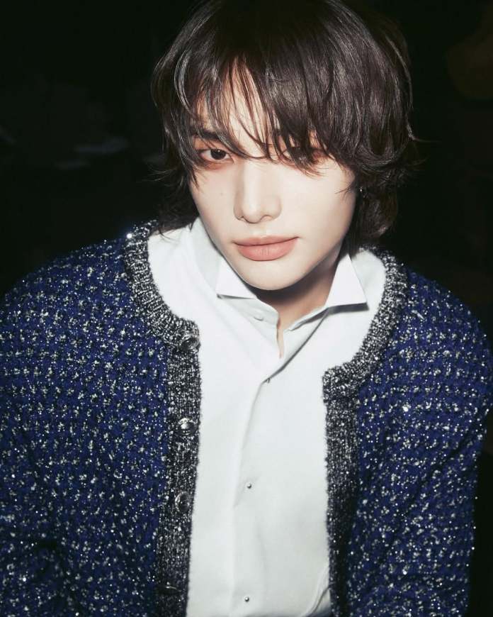 Hyunjin with long, dark hair wearing a blue patterned cardigan over a white shirt, gazing slightly off-camera against a dark background.