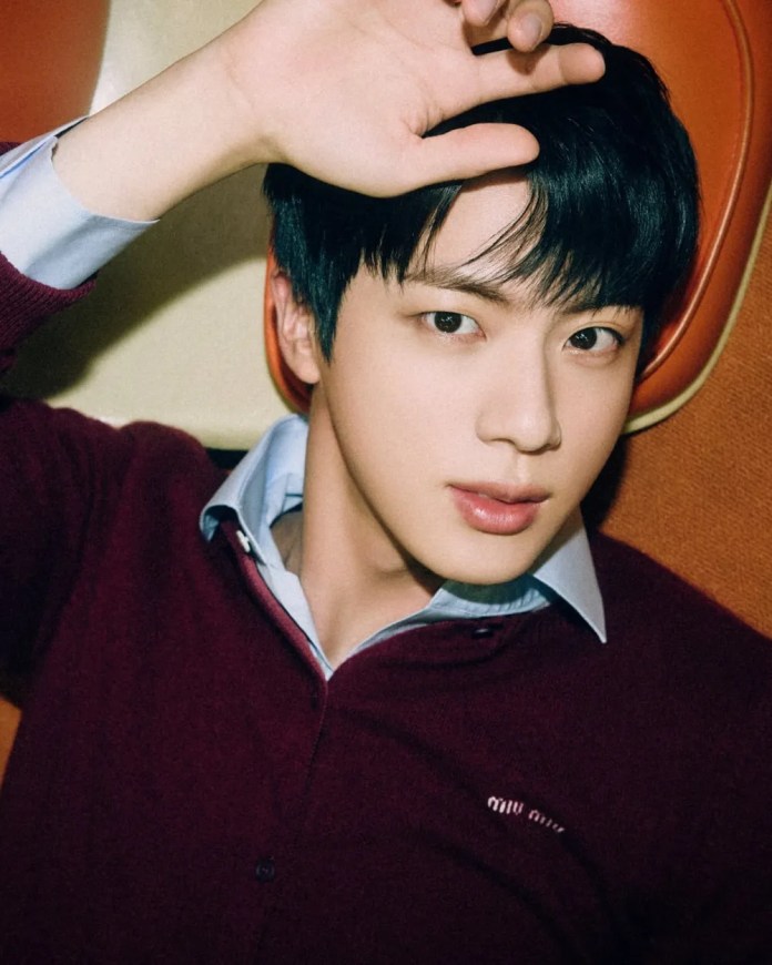 A close-up portrait of a jin with dark hair, wearing a maroon sweater over a light blue shirt, lying down with his hand raised to his forehead against a warm-toned background.