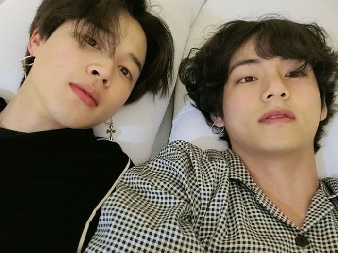 V and Jimin lying on a bed, taking a selfie, with one wearing an earring and both having stylish hairstyles.