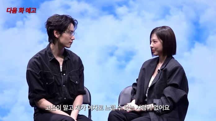 Kim Go eun and Jo Igeon engaged in a conversation on a stage with a cloudy background. One person is wearing glasses and a black shirt, while the other is dressed in a black oversized blazer.