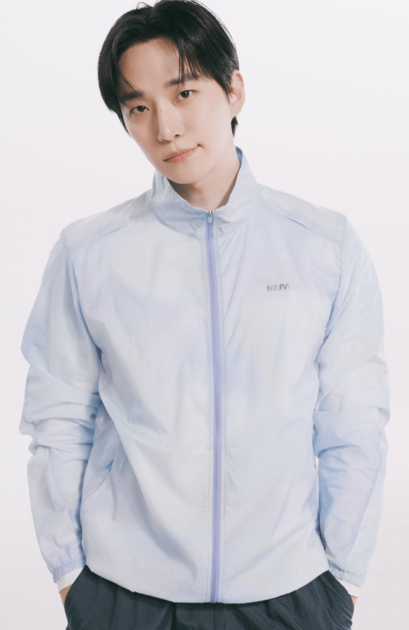 Lee Junho wearing a light blue zip-up jacket and gray pants, standing with his hands in his pockets against a white background.
