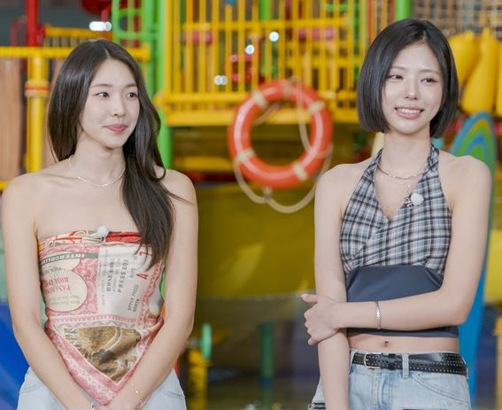 Single's inferno cast standing in front of a colorful playground structure, engaged in conversation. One woman is wearing a strapless top with a print design, while the other is in a sleeveless checked top. A lifebuoy is visible in the background.