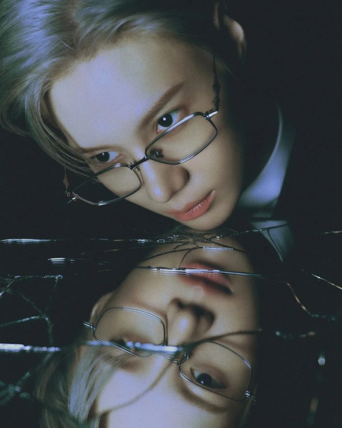 Close-up of taemin wearing glasses, looking intently at their reflection in a broken mirror, with a dark background and soft lighting.