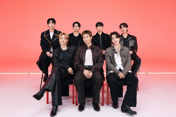 BTS posing for a photo in front of a bright red background. They are dressed in stylish black and neutral outfits, sitting and standing in two rows.