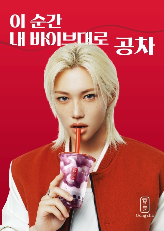 A young person with blonde hair wearing a red and white jacket, holding a cup of purple bubble tea with a red straw. The background is red with Korean text promoting the drink.