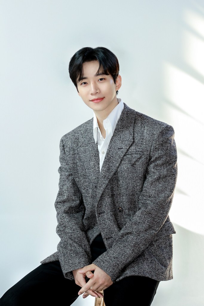 Lee junho with dark hair sits smiling in a stylish grey herringbone blazer and white shirt against a soft, light background.