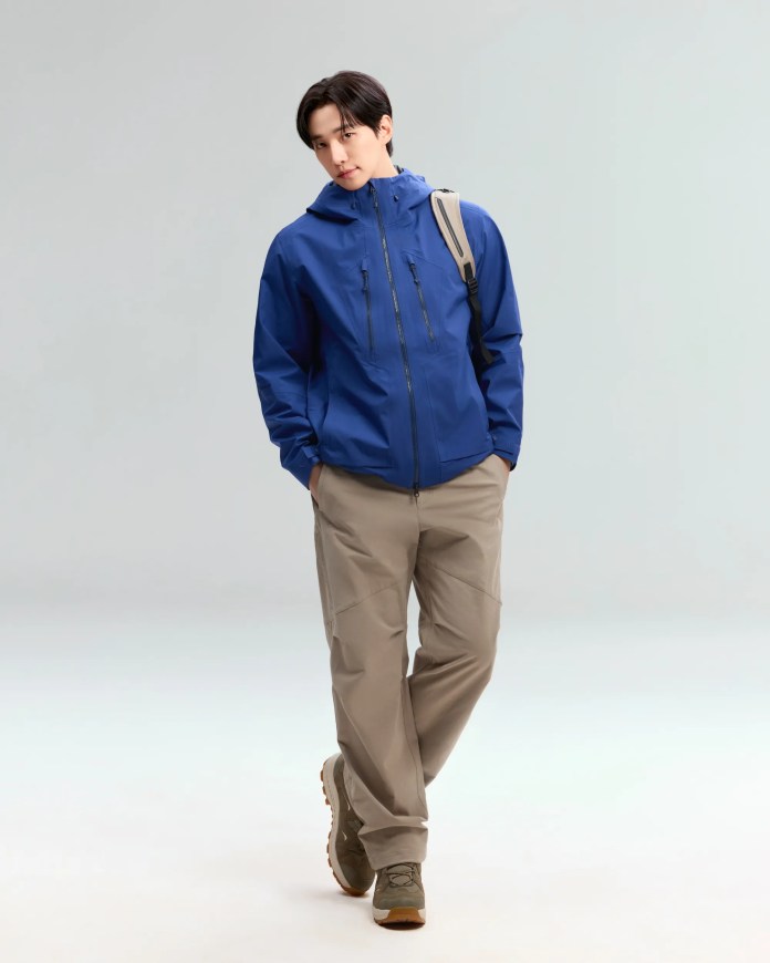 Lee Junho wearing a blue hooded jacket, khaki pants, and green hiking shoes, walking confidently against a neutral background.