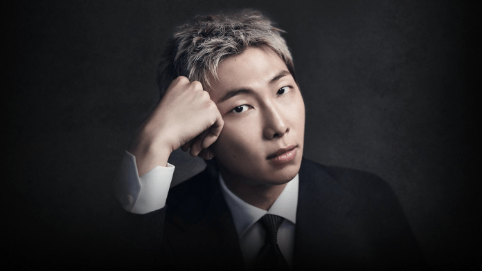 A portrait of RM with short, light-colored hair, wearing a dark suit and tie, posing thoughtfully with his hand resting against his chin against a dark background.