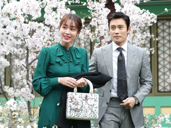 A woman in a green dress and a man in a gray suit are smiling as they walk together among blooming cherry blossom trees.
