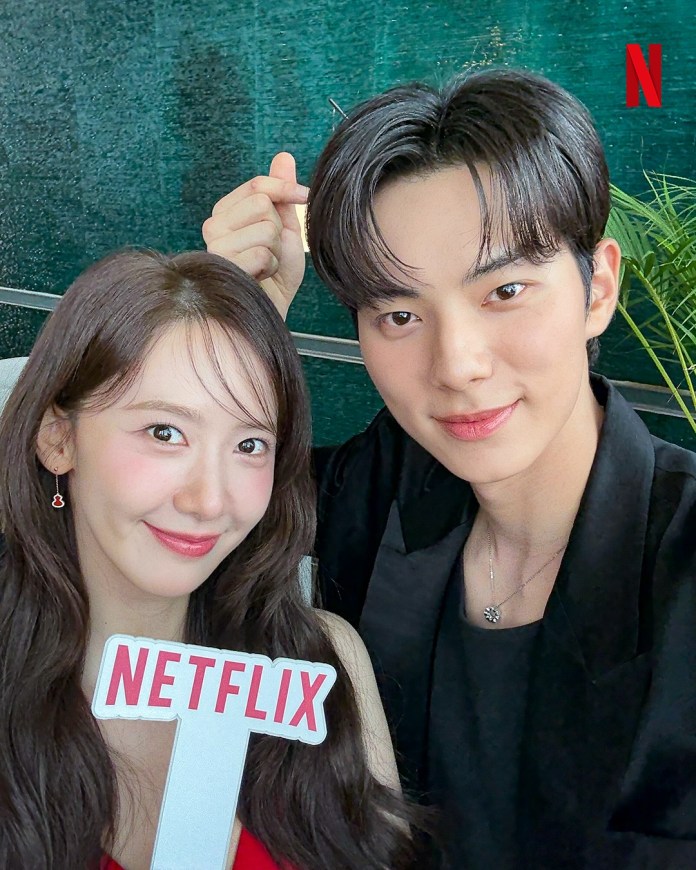A young woman and man smiling together, the woman holding a sign that says 'NETFLIX'. They are posing for a selfie with a green background.