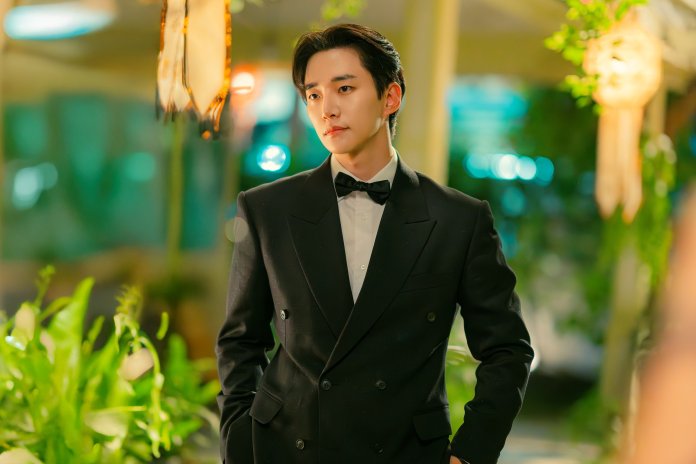 Junho in a black tuxedo with a bow tie, standing in a softly lit outdoor setting adorned with greenery.