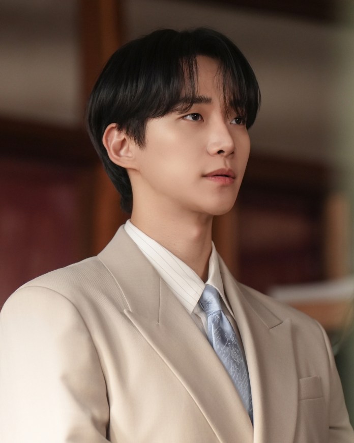 Junho wearing a light beige suit with a white shirt and a light blue tie, looking thoughtfully to the side.