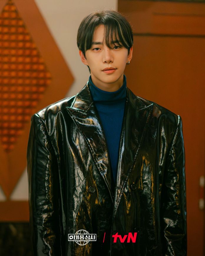 Junho with long hair wearing a shiny black overcoat and a blue turtleneck, standing against a blurred indoor background.