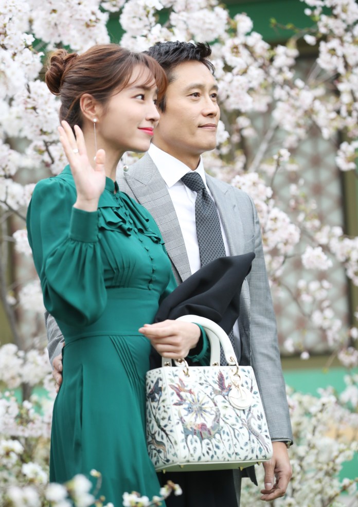 A woman in a green dress waves while posing with a man in a gray suit, set against a backdrop of blooming cherry blossoms.