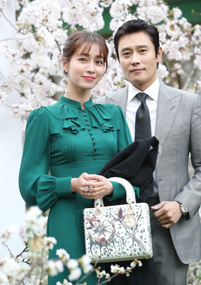 A woman in a green dress and a man in a checked suit pose together in front of blossoming cherry trees, with the woman holding a decorative handbag.