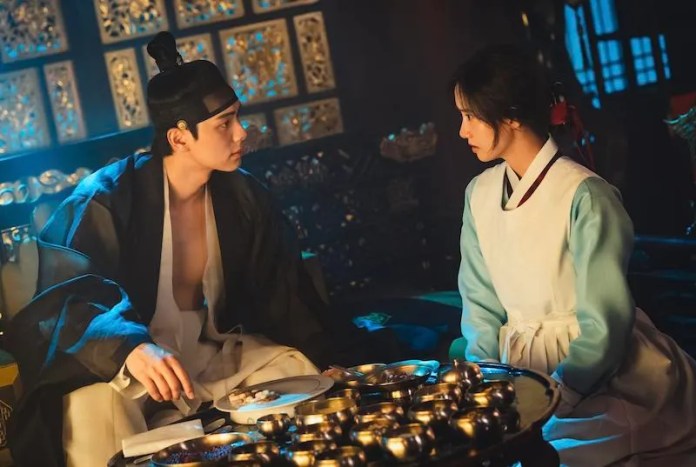 A man and a woman engage in a serious conversation while seated at a low table filled with various dishes in a traditionally decorated room.