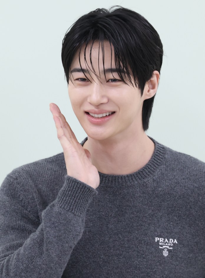 Byeon woo Seok with dark hair is waving his hand in a greeting gesture, wearing a gray sweater with a logo.