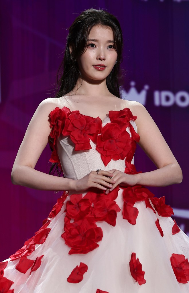 IU wearing a white dress adorned with red flower petals, posing gracefully against a colorful background.