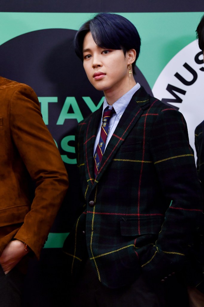 Jimin with dark blue hair, wearing a plaid blazer over a light blue shirt and tie, standing confidently with his hand in his pocket.
