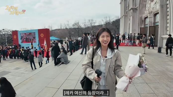 Ha ji won with long hair smiles while holding a bouquet of flowers and a camera, standing on steps in front of a crowd at a festive event. A large screen displays event activities in the background, with flags and people visible.