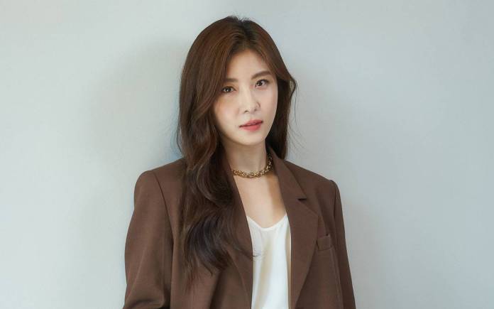  Ha ji won with long brown hair wearing a brown blazer over a white top, standing against a light gray background.