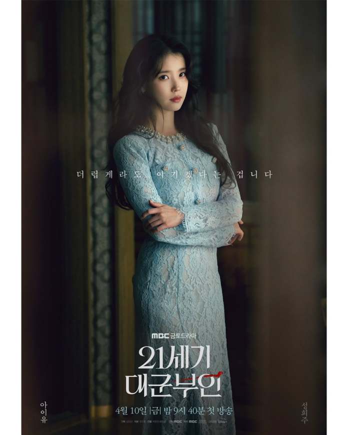 IU in a light blue lace dress poses elegantly with her arms crossed in a dimly lit room, highlighting her serene expression.