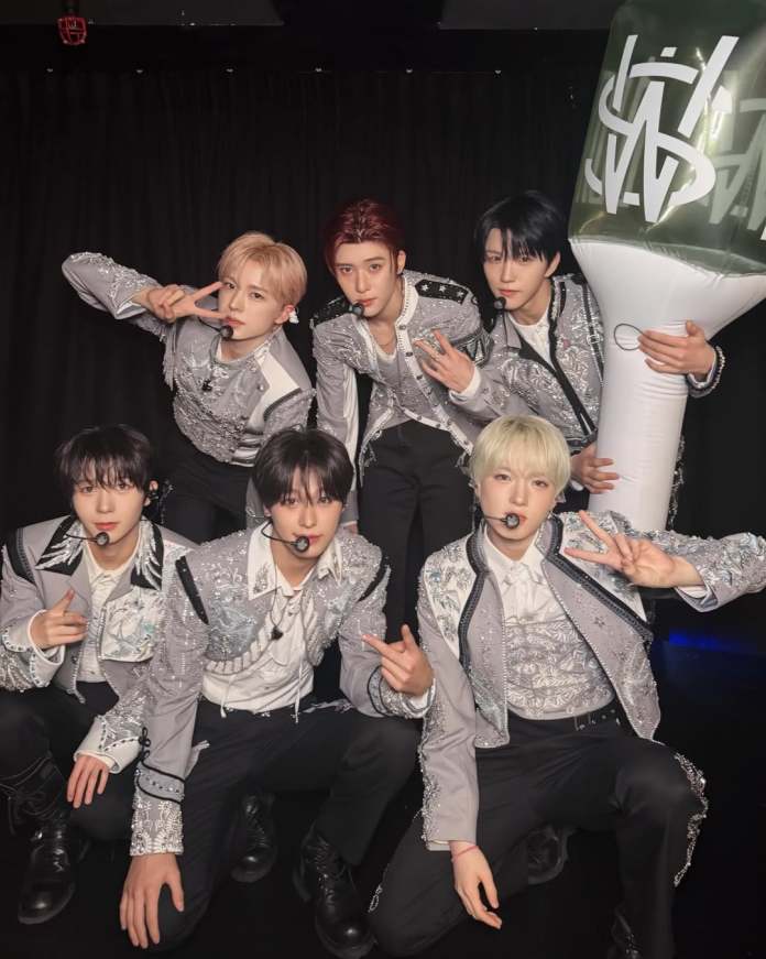 NCT WISH in stylish, embellished outfits posing for a photo, with some making peace signs and others gesturing with hand signs, against a dark background.