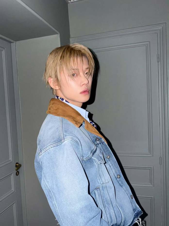 Yeonjun with blonde hair wearing a denim jacket with a brown collar, posed in profile against a gray background.