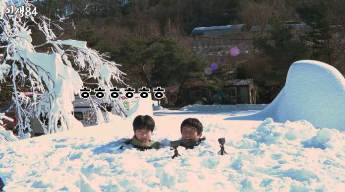 BTS Jin and Kian 84 sitting in a snowy landscape, partially submerged in snow, with trees and a building in the background.