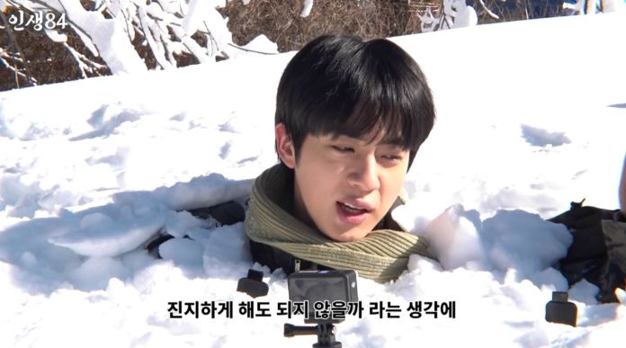 BTS JIN in snow, partially submerged, speaking with a concerned expression. Snowy landscape and trees in the background.