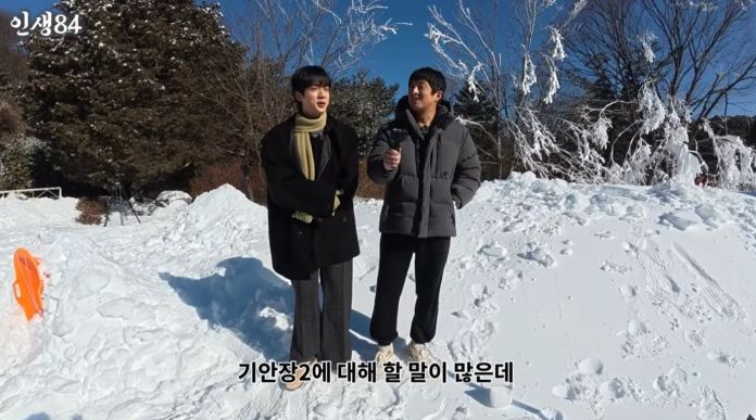 BTS’s Jin and Kian standing in a snowy landscape, with trees and snow-covered ground around them. One person is wearing a long coat with a scarf, while the other is dressed in a puffy jacket, holding a microphone.