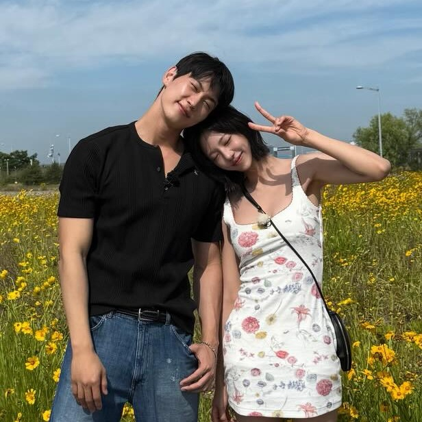 Kim Go Eun and Jo Igeon stands together in a yellow flower field, smiling and posing for the camera. The man, wearing a black shirt and jeans, leans his head toward the woman's shoulder. The woman, in a floral dress, makes a peace sign with her hand.