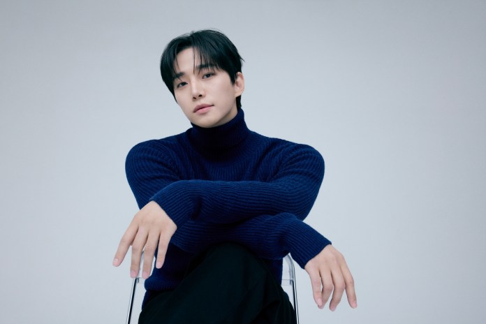 Lee Junho in a dark blue turtleneck sweater poses with his arms crossed while sitting on a clear chair against a light grey background.