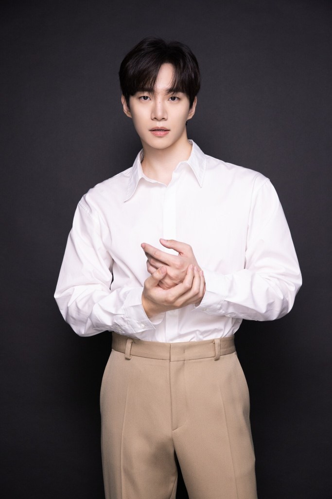 Lee Junho wearing a white shirt and beige pants stands against a dark background, with a serious expression and hands clasped together.