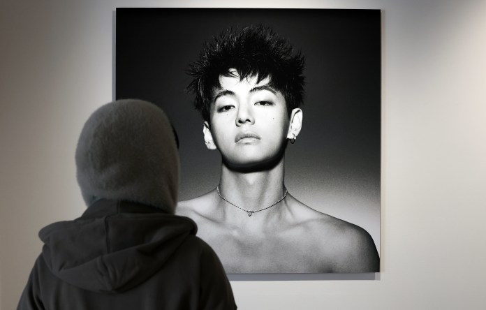 A person wearing a hooded coat stands in front of portrait of a V with a serious expression, short spiky hair, and earrings.