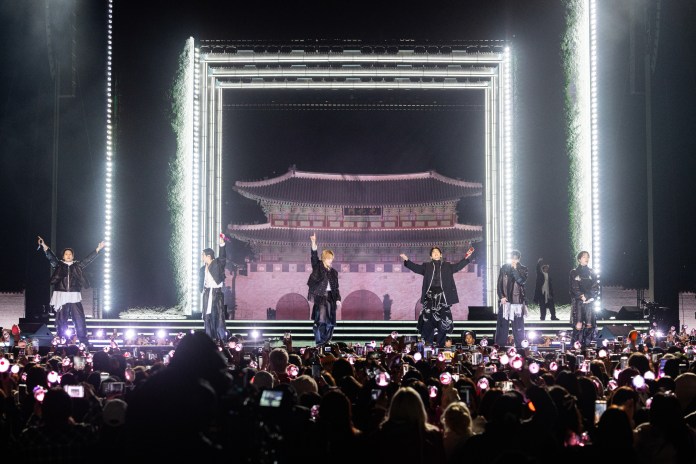 BTS on stage in front of a large audience, illuminated by bright lights and surrounded by fans holding light sticks. Behind them is a traditional architectural structure displayed on a screen.