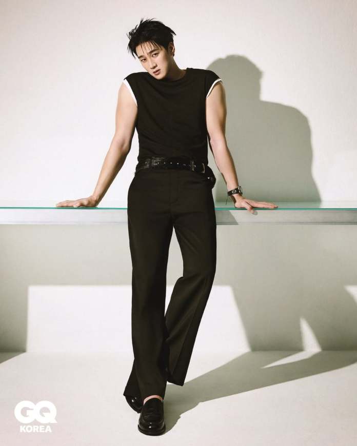 Ahn Bo-hyun posing confidently while leaning on a transparent glass surface, wearing a fitted black sleeveless shirt and black trousers. The background is minimalistic with soft lighting casting shadows.
