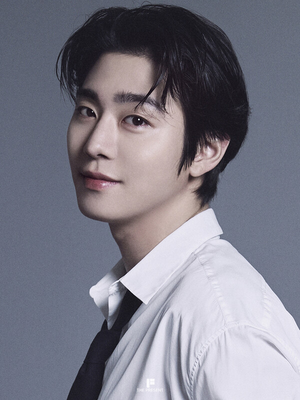 A close-up portrait of Ahn Hyo Seop with dark hair styled slightly tousled, wearing a white shirt and a black tie, smiling at the camera against a gray background.